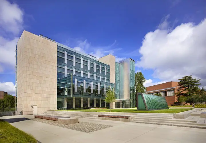 Modern university building with glass facade under blue sky, surrounded by greenery. Sunny day with clouds.