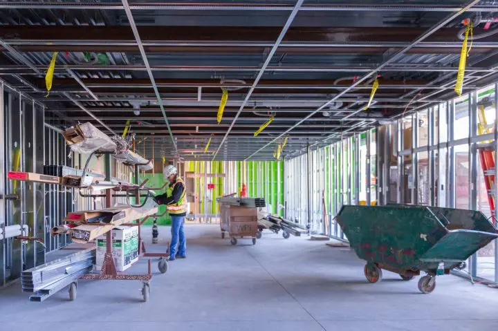 Construction worker in an unfinished building, organizing materials. Safety gear, scaffolding, and green walls visible.