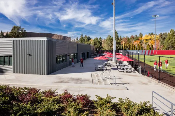Outdoor sports facility with modern building, seating areas, red umbrellas, green field, and people enjoying a sunny day.