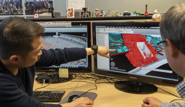 Individuals collaborate on 3D architectural model at dual computer screens, engaging in detailed project discussion.