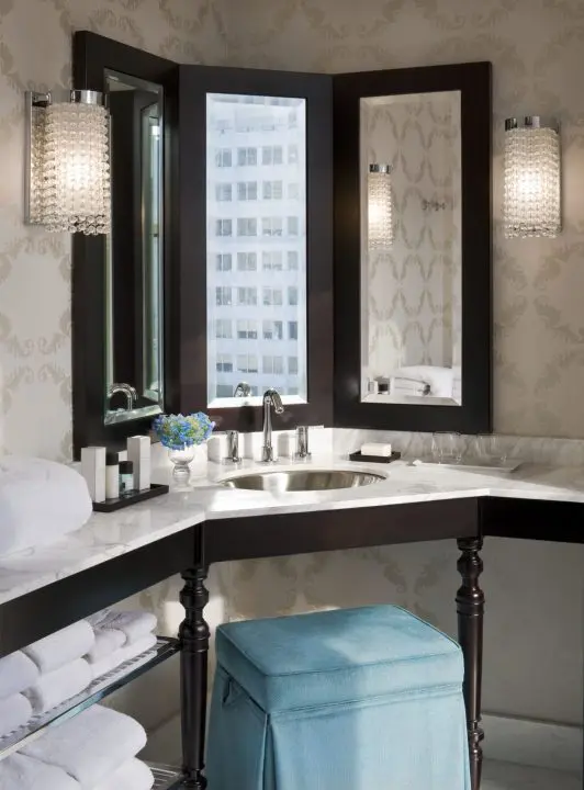 Elegant modern bathroom with corner vanity, soft lighting, and floral decor. Features blue stool and marble countertop.