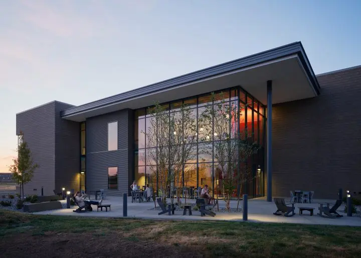 Modern building exterior at dusk with large windows, outdoor seating, and people relaxing, trees subtly lit in the foreground.