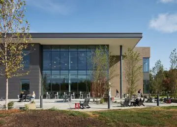 Modern building exterior with large windows, patio seating, and surrounding greenery under a clear blue sky.