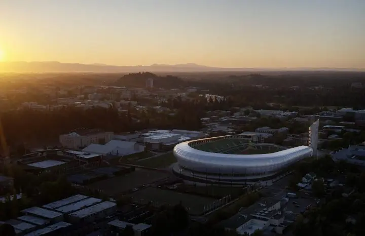 Sunset over cityscape with modern stadium in foreground, surrounded by lush greenery and urban buildings. Scenic evening view.