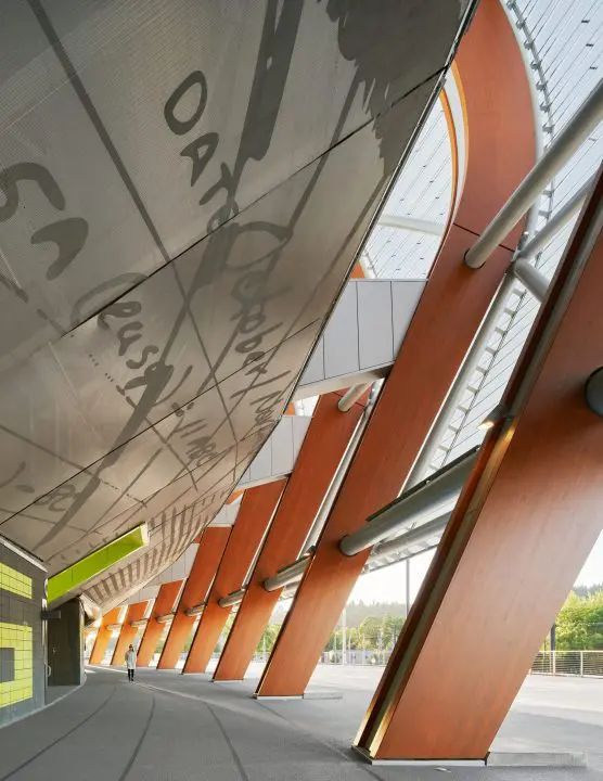 Modern architectural walkway with tall wooden beams and reflective surfaces. A person walks along, highlighting the structure's scale.