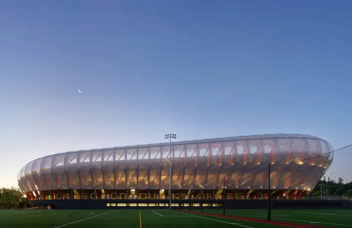 Modern sports stadium with illuminated exterior at twilight, set against a clear sky with a crescent moon.