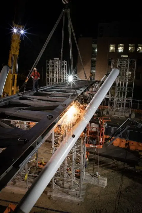 Construction site at night with workers welding a large metal structure, sparks flying, illuminated by lights. Urban development.