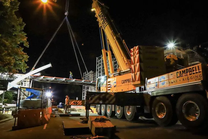 Night construction scene with crane lifting materials onto a bridge. Workers oversee the operation under bright lights.