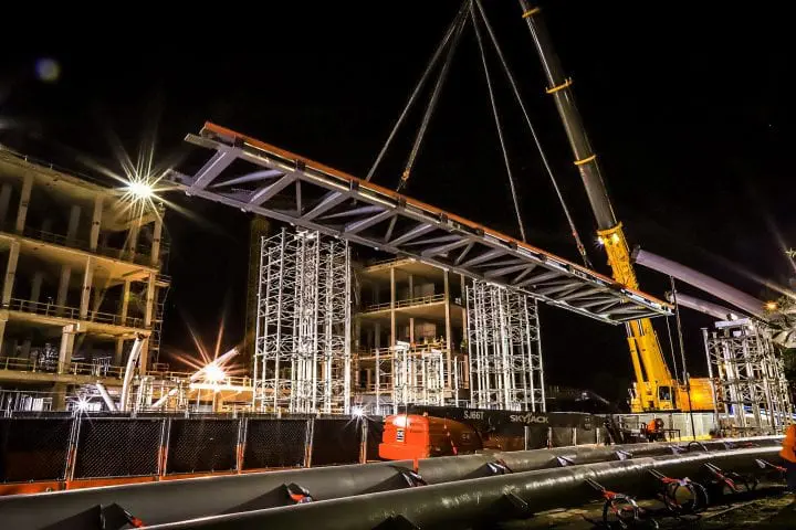 Construction site at night with a crane lifting large steel beams, illuminated by bright lights, against dark sky.