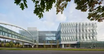 Modern glass building with geometric design and connecting bridge, framed by trees against a blue sky. Road in foreground.