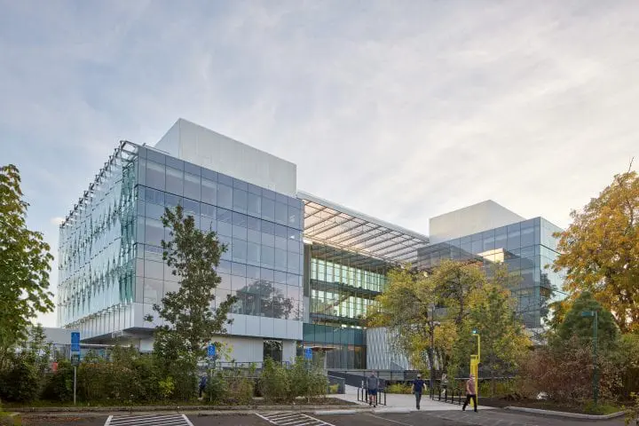 Modern glass building with trees and people walking in the foreground, under a clear sky.