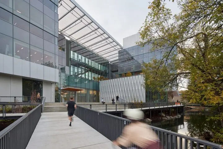 Modern glass building with people walking and cycling on a bridge, surrounded by trees, near a peaceful water body.