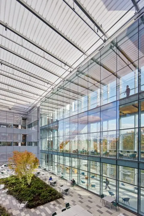Modern glass-walled building with open courtyard, greenery, and seating. Bright natural light and spacious interior visible.