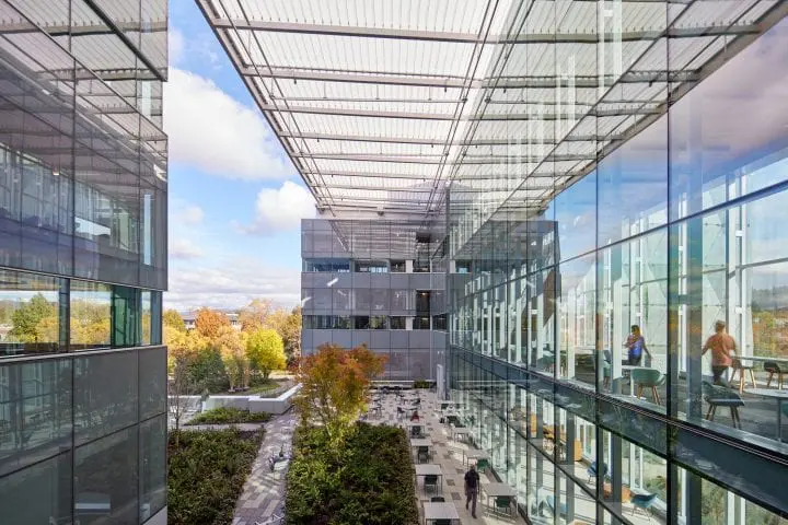 Modern office building with glass facades and outdoor terrace seating surrounded by trees under a blue sky.