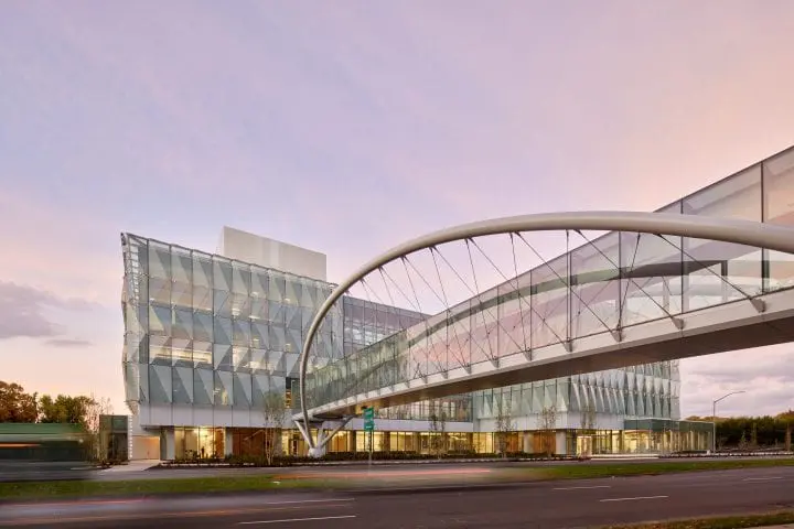 Modern glass building with curved sky bridge, under a pink sunset. Urban architecture and design elements.