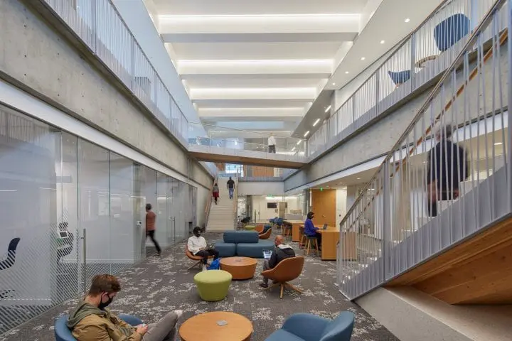 Modern open space with people working and socializing, featuring concrete, glass walls, and stylish furniture in a spacious building lobby.