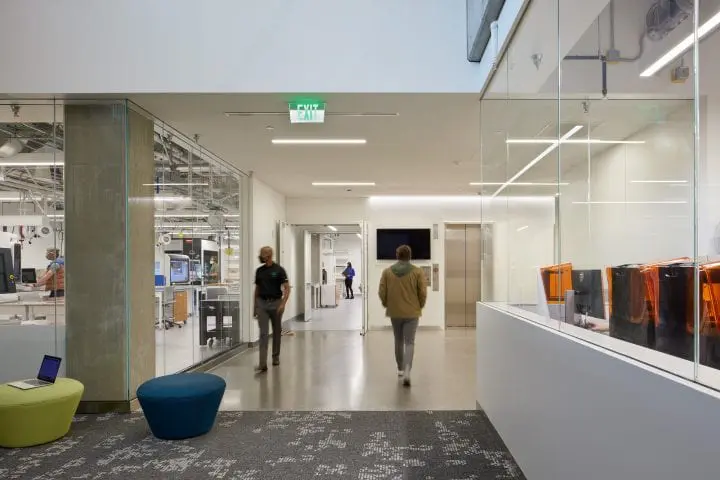 Modern office space with individuals walking and working, featuring glass walls, office equipment, and casual seating.