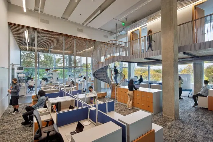 Modern open office with spiral staircase, collaborative workspaces, and people engaged in discussions and tasks.
