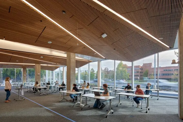 Modern classroom with individuals at desks, observing distancing guidelines, large windows, and wooden ceiling design.
