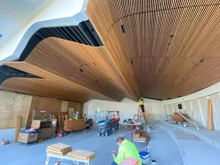 Construction workers install curved wooden ceiling panels in a spacious room. Scaffolding, tools, and materials are visible.