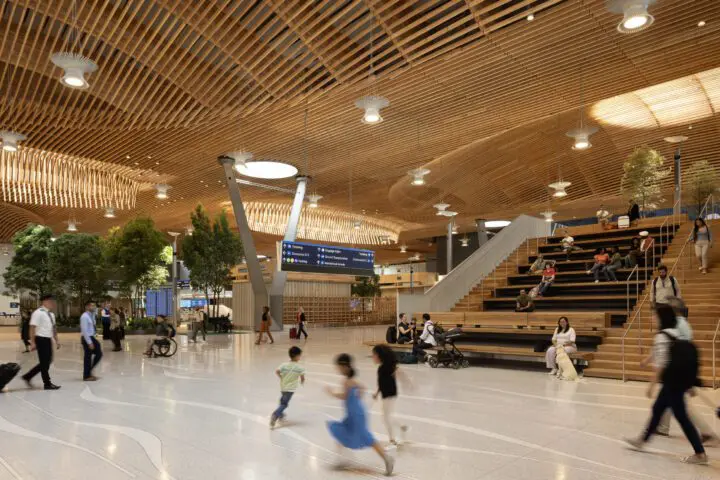 Spacious airport terminal with wooden ceilings, travelers walking, sitting areas with greenery, and directional signs.