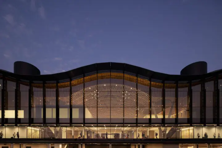 Modern architectural building with glass facade, illuminated at dusk. Silhouettes of people walking inside, under a twilight sky.