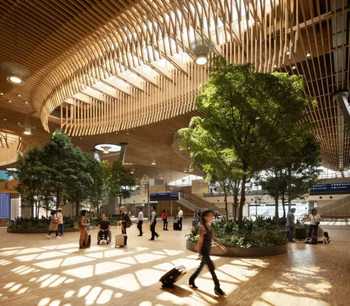 Modern airport terminal with wood ceiling, natural light, and travelers walking past indoor trees and plants.