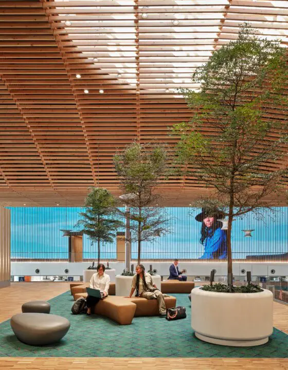 Modern airport lounge with greenery, gender-neutral seating, and people using laptops under a wooden ceiling with a large screen display.