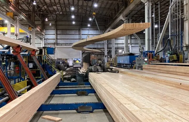 Wood manufacturing facility with large timber beams in production process. Industrial setting with machinery and assembly lines.