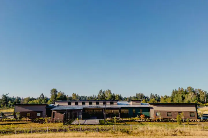 Modern building with flat roof in a rural landscape, surrounded by greenery and trees under a clear blue sky.
