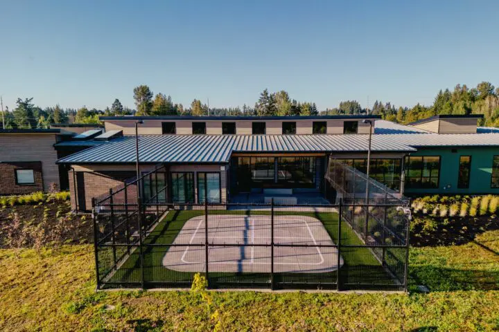 Modern building with metal roof, featuring a fenced outdoor sports court, surrounded by lush greenery and trees under a clear blue sky.