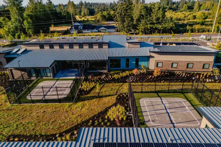 Aerial view of a modern building with solar panels and sports courts, surrounded by greenery and forested background.
