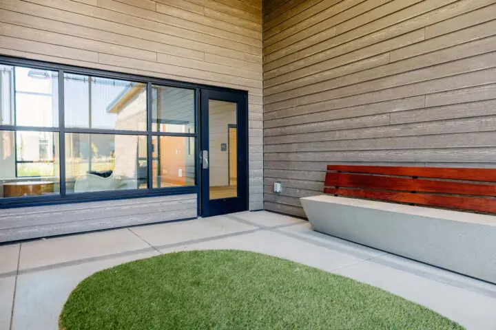 Modern building entrance with a large window, door, and a wooden bench on a concrete base beside a grass patch.
