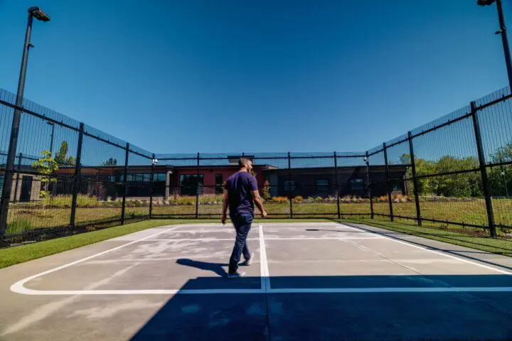 Person walking on an outdoor court, surrounded by a tall fence and greenery. Clear blue sky overhead.