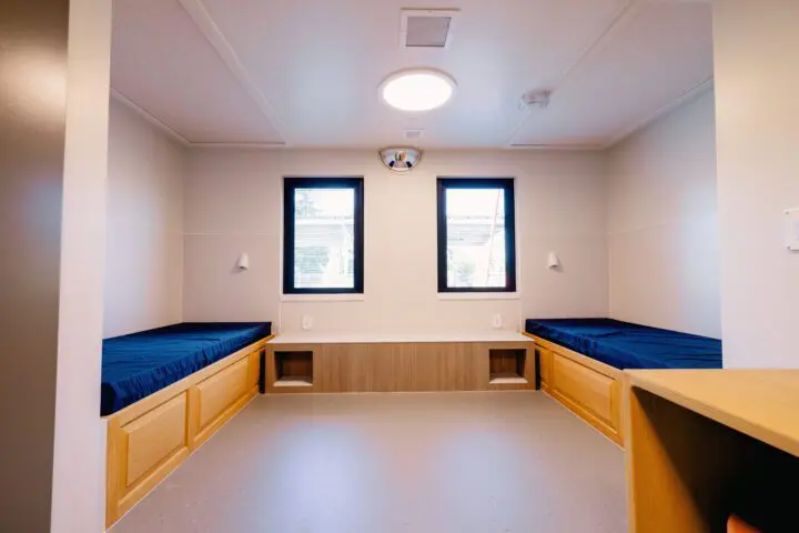 Minimalist room with twin beds, blue bedding, natural light from two windows, and simple wood furnishings.