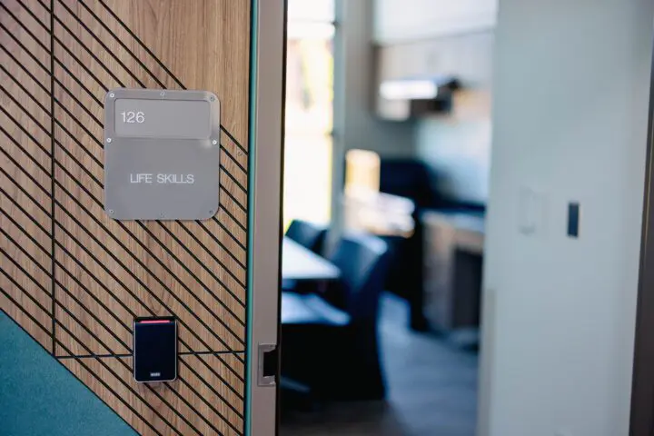 Entrance to Life Skills room 126 with modern wood paneling and card reader, leading to a bright interior space with tables and chairs.
