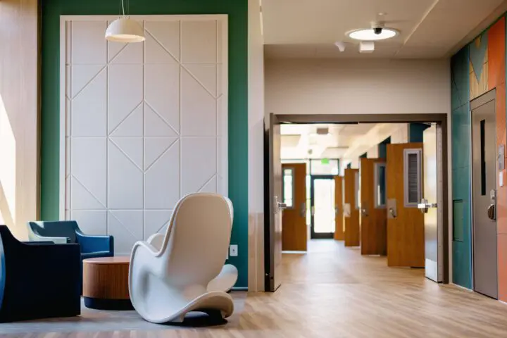 Modern waiting area with sleek chairs, wooden table, and geometric wall design next to a bright hallway.