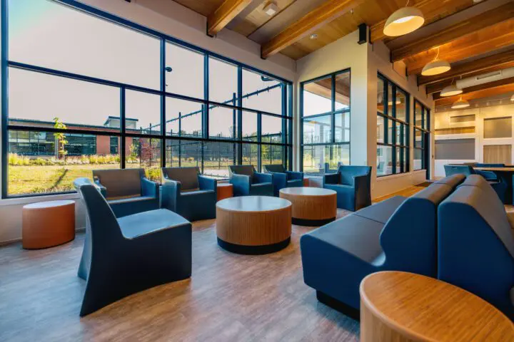 Modern lounge area with blue seating and round wooden tables, large windows showcasing outdoor greenery. Bright and inviting interior space.