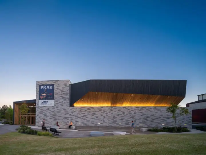 Modern building with a bold design at dusk, featuring a illuminated wooden canopy and a poster promoting an event.