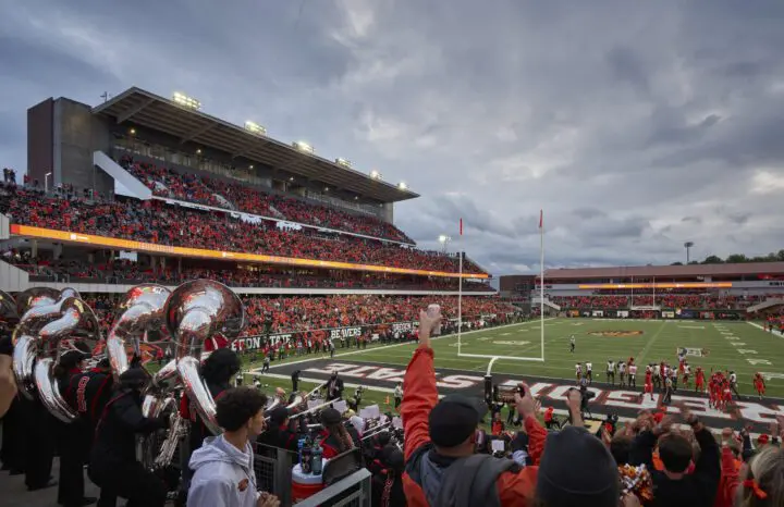 Vibrant football game scene with a marching band, cheering crowd, and players on the field under a cloudy sky.