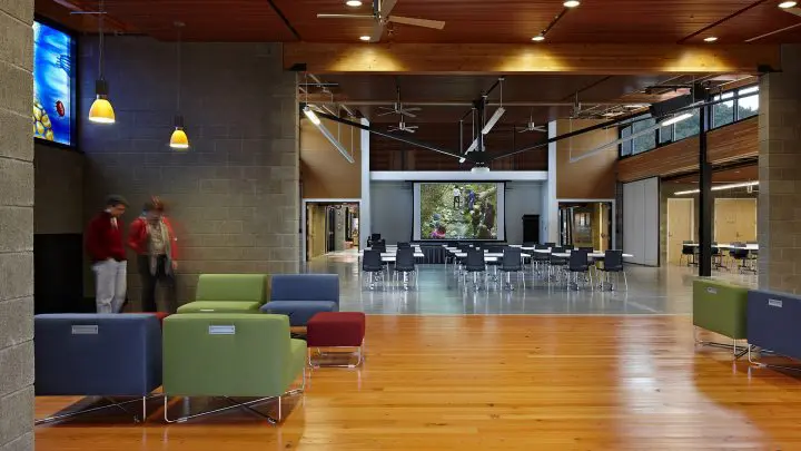 Modern educational facility with seating, projector, and open layout. Two individuals in motion. Bright and spacious atmosphere.