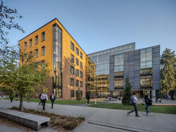 Modern university building with glass windows and a brick facade, surrounded by people walking on campus under a clear blue sky.