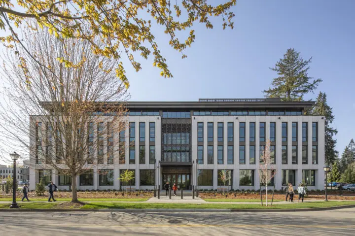 Modern building with large windows, surrounded by trees. People walk along the path on a sunny day.