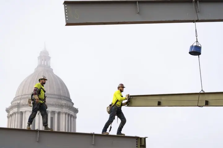Construction workers on steel beams near a domed building, city skyline in the background. Safety gear and teamwork in action.