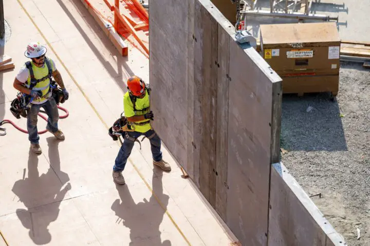 Construction workers guide a concrete wall panel into place at a building site, wearing safety gear and vests.