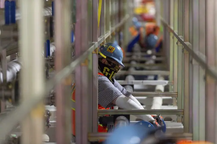 Worker in safety gear inspecting pipes in an industrial setting, focusing on machinery maintenance and safety protocols.