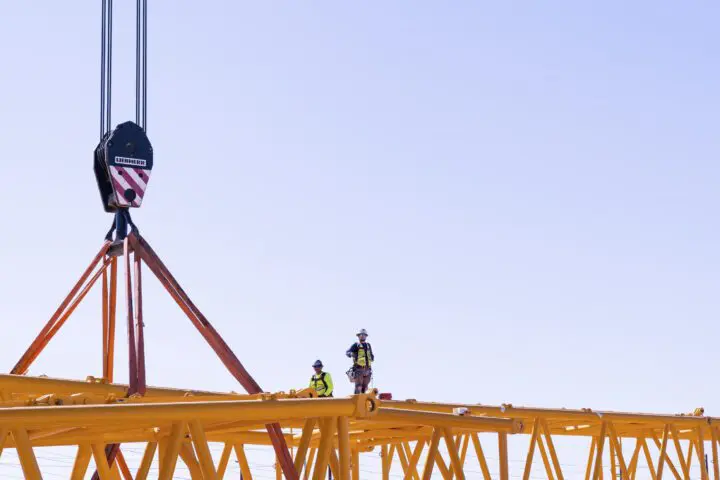 Construction crew working on yellow crane assembly against clear sky. Safety gear visible.