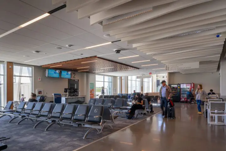 Airport waiting area with seating, vending machine, and people relaxing. Bright, modern interior with clear signage to baggage claim.