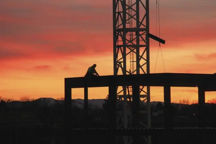 Silhouette of a lone construction worker on a steel frame at sunset, with orange skies highlighting the industrial scene.