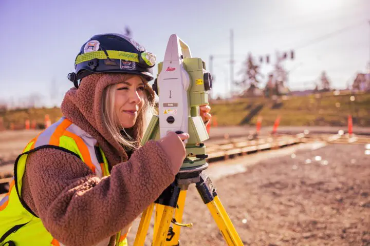 A construction worker uses a theodolite on a sunny job site, wearing safety gear and a hard hat.